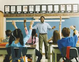 A teacher stands at the front of a classroom engaging with students who are seated at desks and eagerly raising their hands. The students are diverse in appearance, and the classroom setting includes a chalkboard, framed artwork above it, and typical school furniture, illustrating an interactive and inclusive learning environment.